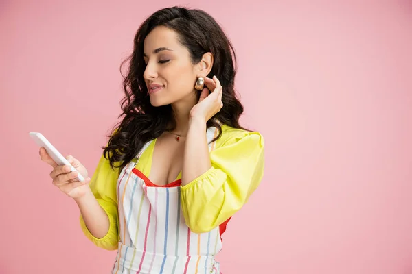 Pleased housewife in striped apron touching wavy hair while looking at mobile phone isolated on pink — Stock Photo