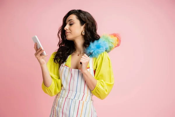 Casalinga scontento in grembiule a righe tenendo pennello polvere colorato e guardando il telefono cellulare isolato su rosa — Foto stock