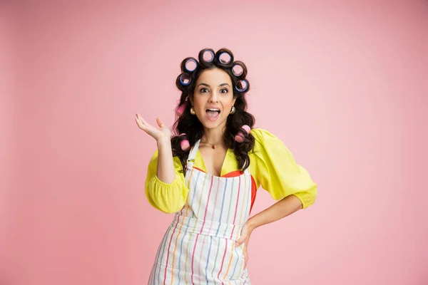 Dona de casa espantada em avental listrado e encrespadores de cabelo de pé com a mão no quadril isolado em rosa — Fotografia de Stock