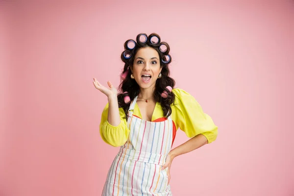 Amazed housewife in striped apron and hair curlers standing with hand on hip isolated on pink — Stock Photo