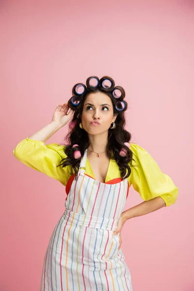 Dona de casa pensativa em avental listrado e blusa amarela tocando encrespadores de cabelo e olhando para longe isolado em rosa — Fotografia de Stock