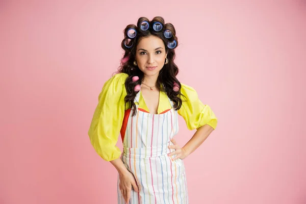 Young brunette housewife in hair curlers and striped apron on yellow blouse looking at camera isolated on pink — Stock Photo