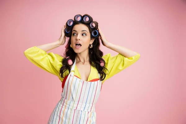 Amazed and curious housewife in striped apron adjusting hair curlers and looking away isolated on pink — Stock Photo