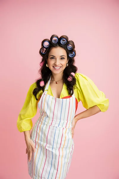 Positive woman in hair curlers and striped apron holding hand on hip and smiling at camera isolated on pink — Stock Photo