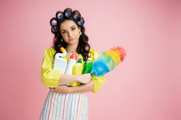 Bored housewife with hair curlers and different cleaning supplies looking at camera isolated on pink — Stock Photo