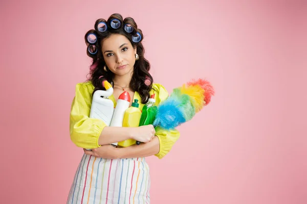 Gelangweilte Hausfrau mit Lockenwicklern und verschiedenen Putzutensilien schaut vereinzelt in die Kamera auf rosa — Stockfoto