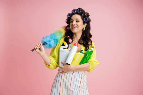 Cheerful housewife in hair curlers holding colorful feather duster and cleaning supplies while looking away isolated on pink — Stock Photo