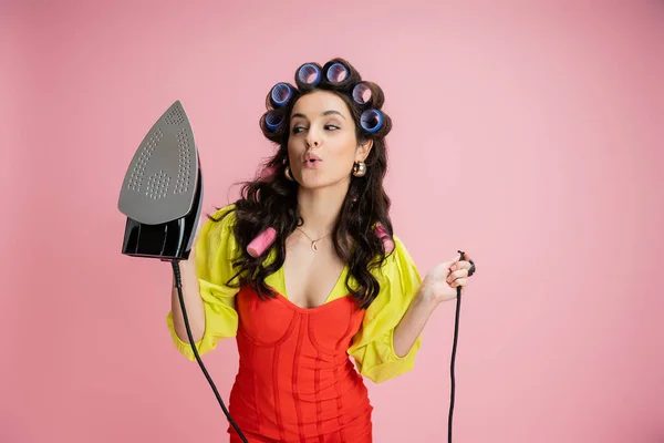 Housewife in hair curlers and yellow blouse with red corset dress looking at iron isolated on pink — Stock Photo