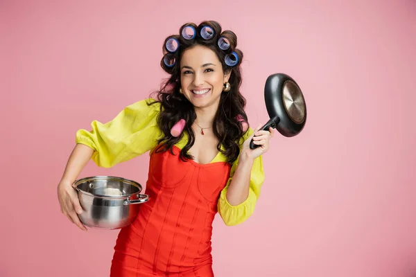 Веселая домохозяйка в красном корсетном платье и бигуди для волос держа кастрюлю и сковородку, глядя на камеру, изолированную на розовый — стоковое фото