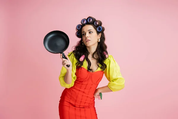 Bored housewife in hair curlers and red corset dress looking at frying pan isolated on pink — Stock Photo