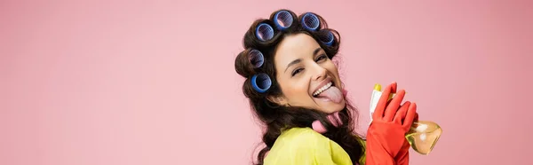 Flirty housewife in hair curlers and red rubber gloves sticking out tongue while holding spray bottle isolated on pink, banner — Stock Photo
