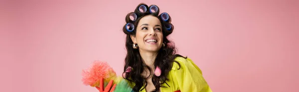 Dona de casa alegre em encrespadores de cabelo segurando escova de poeira e olhando para longe isolado em rosa, banner — Fotografia de Stock