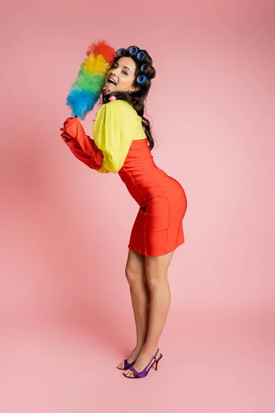 Full length of flirty housewife in red corset dress and hair curlers posing with colorful dust brush on pink — Stock Photo
