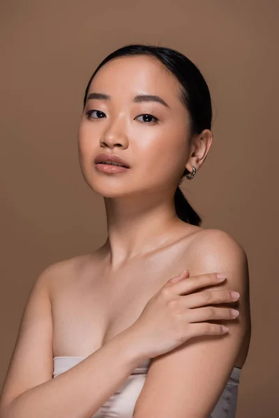 Young asian woman in top touching shoulder and looking at camera isolated on brown — Foto stock