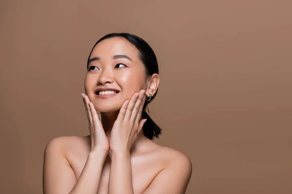 Cheerful asian model with naked shoulders touching cheeks isolated on brown — Foto stock