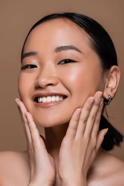 Portrait of young asian woman with naked shoulders touching cheeks isolated on brown — Stock Photo