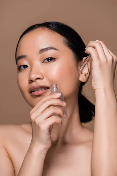 Asian woman with naked shoulders holding serum isolated on brown — Stock Photo