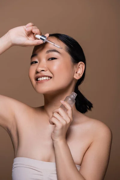 Pleased asian woman in top holding pipette and bottle of cosmetic serum isolated on brown — Stock Photo