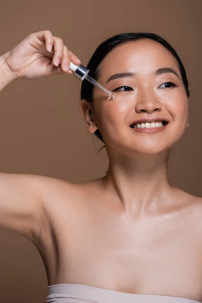 Smiling asian woman applying serum on face isolated on brown — Stock Photo