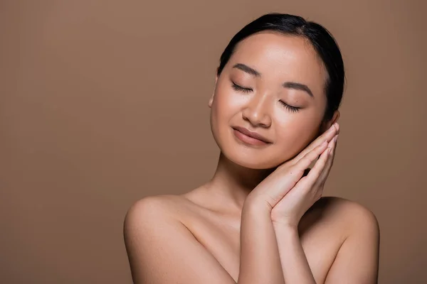 Young brunette asian woman with naked shoulders holding hands near cheek isolated on brown — Stock Photo