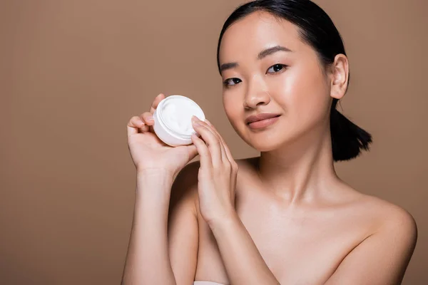 Pretty asian woman with naked shoulders holding container with cosmetic cream isolated on brown — Photo de stock
