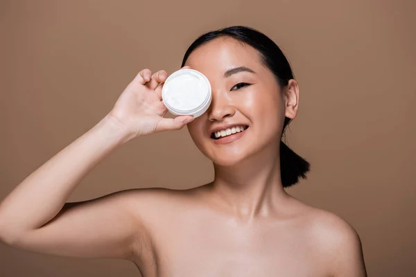 Positive asian woman with naked shoulders holding container with cosmetic cream isolated on brown — Stock Photo