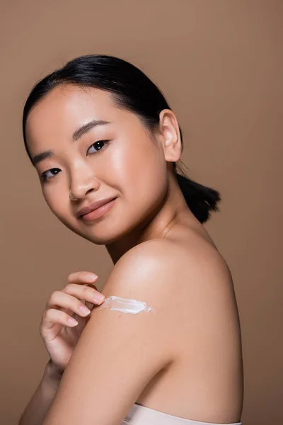 Smiling asian woman applying cosmetic cream on shoulder isolated on brown — Photo de stock