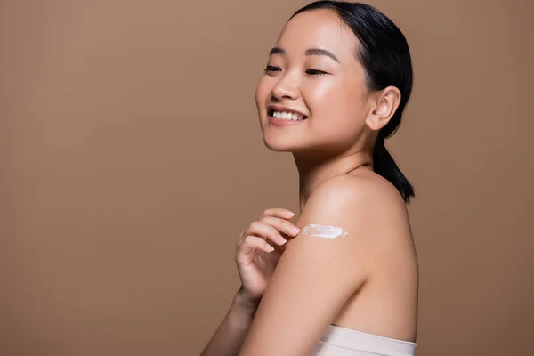 Cheerful asian model applying cosmetic cream on shoulder isolated on brown — Foto stock