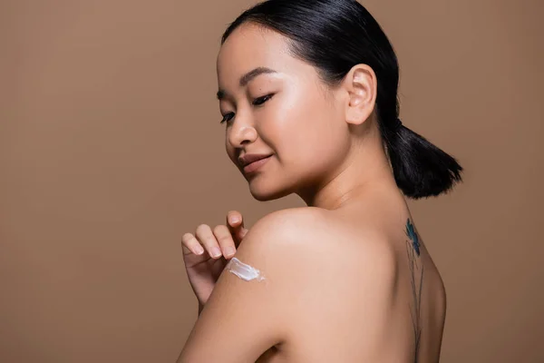 Татуйована азіатка з голими плечима наносить косметичний крем на плече ізольовані на коричневому — стокове фото