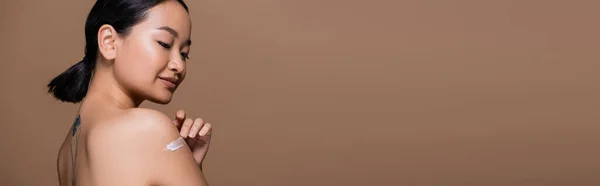 Татуйована азіатська модель нанесення косметичного крему на плече ізольовано на коричневому, банер — стокове фото