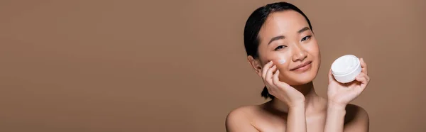 Smiling asian model applying face cream on cheek isolated on brown, banner — Stock Photo