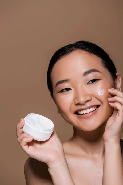 Positive asian woman with naked shoulders applying face cream isolated on brown — Stock Photo
