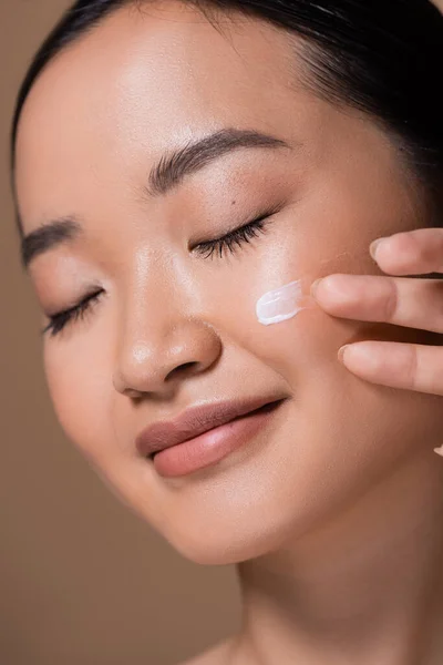 Close up view of pretty asian woman applying face cream and closing eyes isolated on brown — Stock Photo