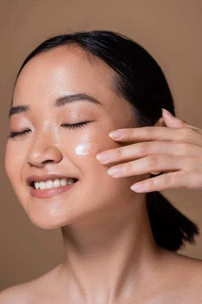 Smiling asian woman applying face cream isolated on brown — Photo de stock