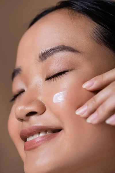 Close up view of young asian woman with face cream on cheek isolated on brown — Fotografia de Stock