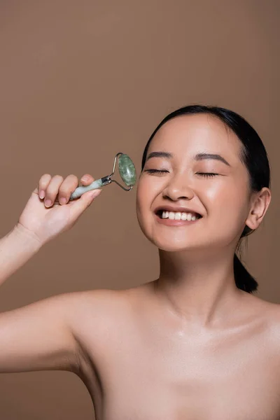 Smiling asian woman with naked shoulders holding jade roller isolated on brown — Fotografia de Stock