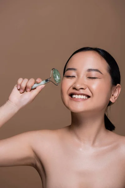 Smiling asian woman with naked shoulders holding jade roller isolated on brown — Stock Photo