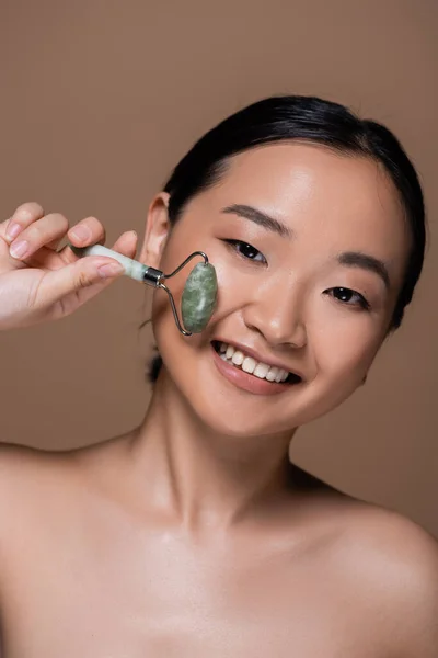Весела азіатська жінка з голими плечима тримає нефритовий ролик ізольовано на коричневому — стокове фото