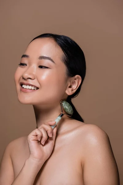 Smiling young asian woman with naked shoulders holding jade roller isolated on brown — Stock Photo