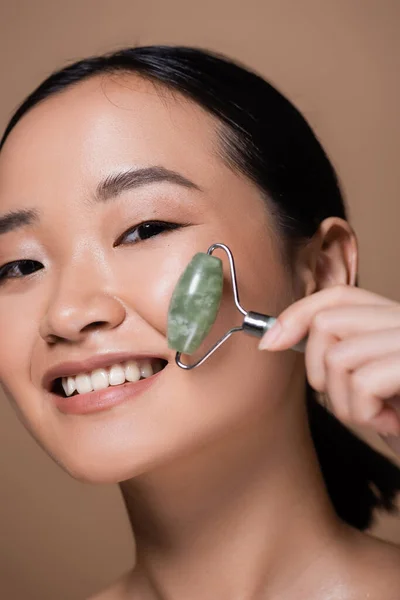 Portrait of cheerful asian woman holding jade roller and looking at camera isolated on brown - foto de stock