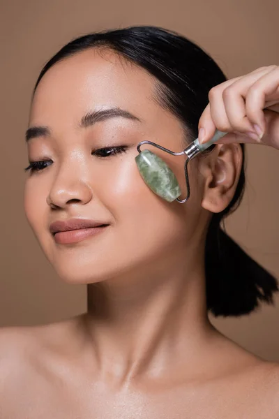 Pretty asian model using jade roller on face isolated on brown — Stock Photo