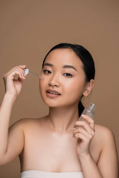 Brunette asian woman applying serum and looking at camera isolated on brown - foto de stock