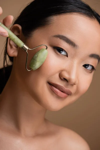 Portrait of brunette asian woman using jade roller on face isolated on brown — Stock Photo