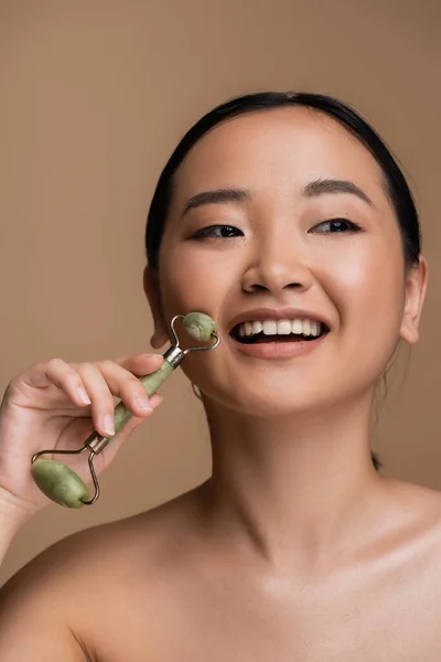 Positive asian model with naked shoulder using jade roller isolated on brown — Stock Photo