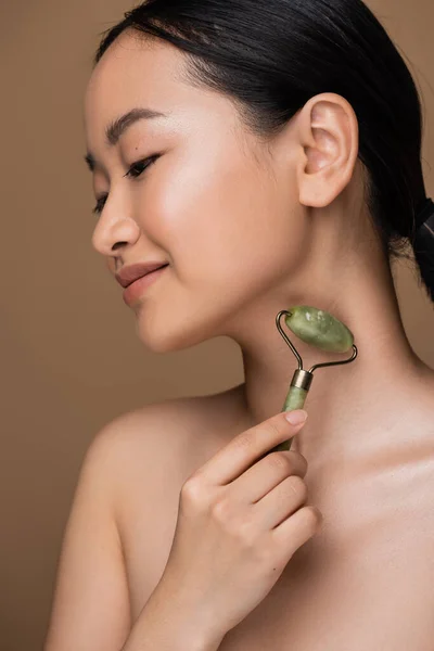 Asian woman with naked shoulder using jade roller on neck isolated on brown — Stock Photo