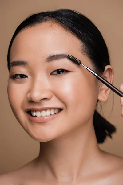 Pretty asian model styling eyebrow while holding brush isolated on brown — Stockfoto