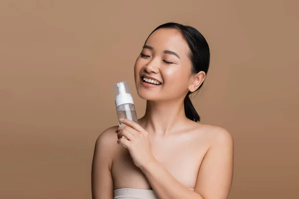 Positive asian model with naked shoulders holding cleansing foam isolated on brown — Photo de stock