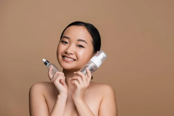 Cheerful asian model holding serum and face foam isolated on brown — Stock Photo
