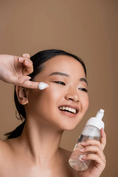 Cheerful asian woman with naked shoulders holding bottle of face foam isolated on brown — Stock Photo