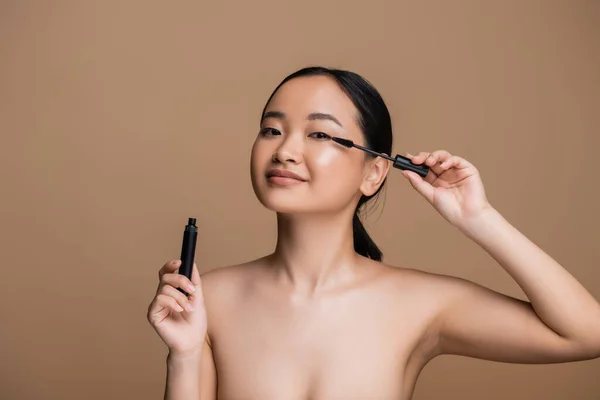 Young asian woman with naked shoulders holding mascara isolated on brown — Stock Photo